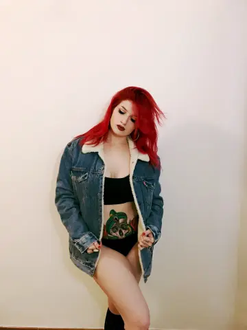 Red-haired woman in a denim jacket and black crop top posing confidently against a plain white wall.