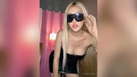 Blonde woman in a black velvet top and sunglasses posing in a pink room with a glamorous aesthetic.