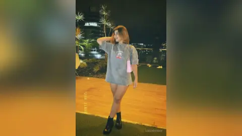 Smiling young woman in a grey oversized graphic t-shirt and black boots relaxing on a rooftop garden at night.