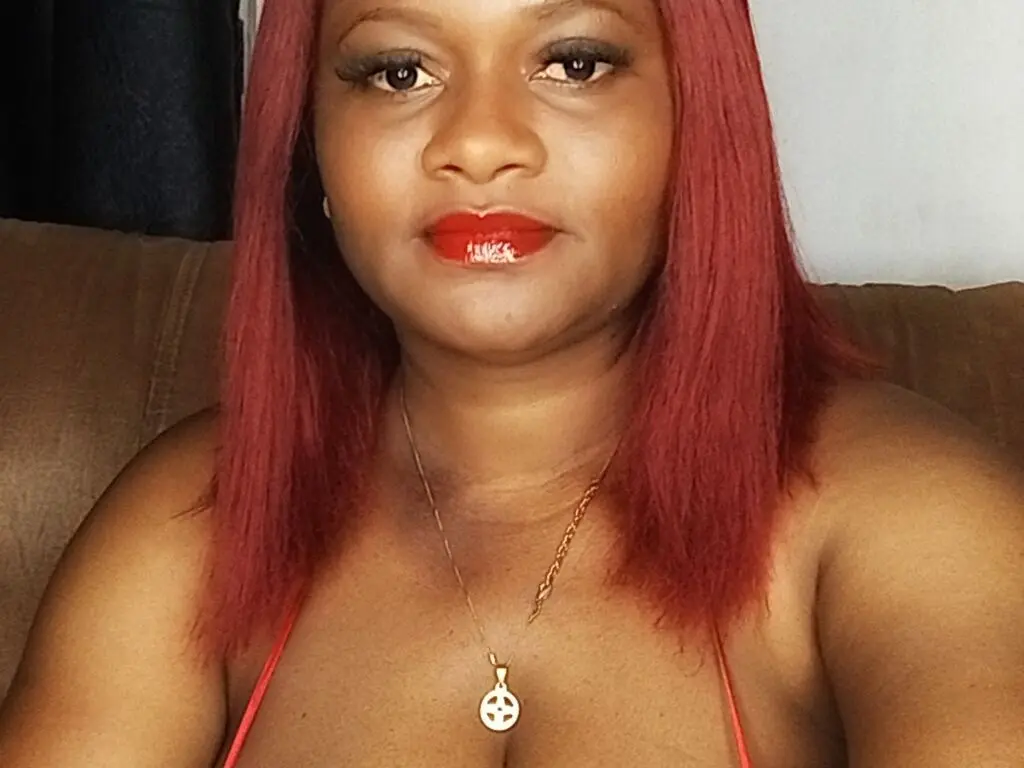 Red-haired woman in a red top wearing a gold pendant necklace indoors with a warm, confident mood.