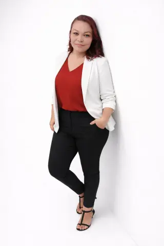 Confident woman in a white blazer, red blouse, and black trousers posing against a white studio background.