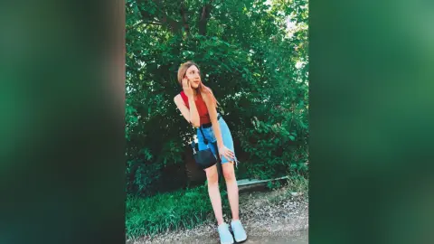 Young woman in a red tank top and denim shorts standing by lush green trees in a sunny outdoor setting.