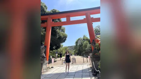Young woman in a black summer dress standing under a traditional red Japanese torii gate on a sunny day.