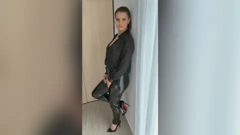 Elegant woman in a black button-down shirt and shiny leather pants posing in a modern minimalist room.