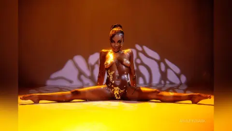 Nude Black woman with gold body paint performing a split in warm studio lighting.