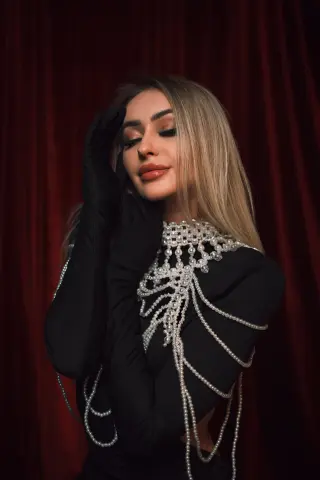 Elegant woman in a black long-sleeve dress with pearl embellishments posing against a red velvet curtain.