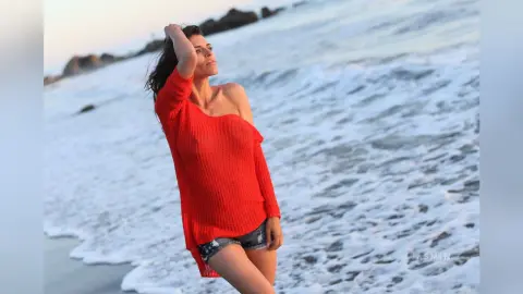 Cheerful woman in a bright red oversized sweater posing on a breezy beach by the ocean waves.
