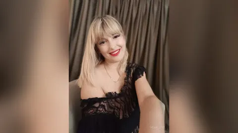 VeraLuxe — VeraLuxe smiles in a black lace top while sitting in front of brown curtains.