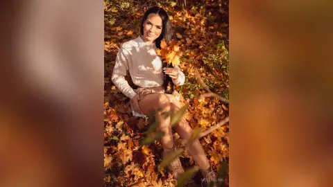 Smiling woman in a white sweater sitting in autumn leaves holding a golden maple leaf in a sunny forest.