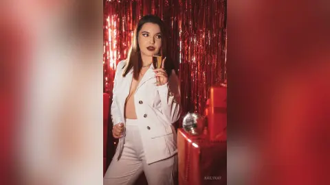 Glamorous woman in a white suit holding a champagne glass against a festive red tinsel background.