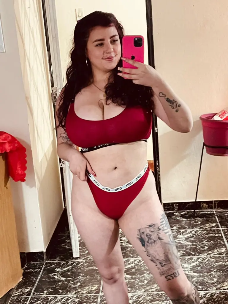 AngelaCutee takes a mirror selfie wearing a low-cut black tank top, showing off her cleavage and long brown hair.