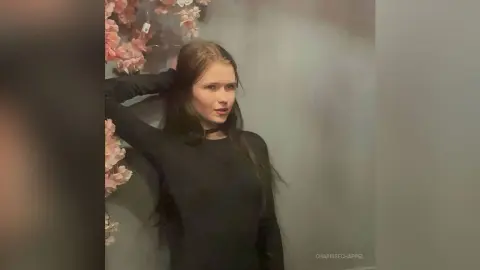 Young woman in a black long-sleeve top posing against a floral wall in a moody, soft-lit setting.