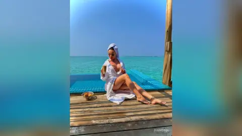 Relaxed woman in a white bathrobe enjoying a drink on a wooden pier overlooking the turquoise ocean.