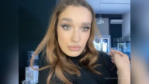 EmelyMastorovo poses for a close-up selfie wearing a black top with her long brown hair styled in loose waves.