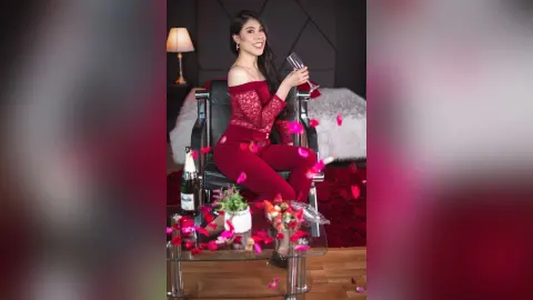 SamanthaHarvey sits in a black chair wearing a red lace off-the-shoulder bodysuit, holding a glass of red wine.