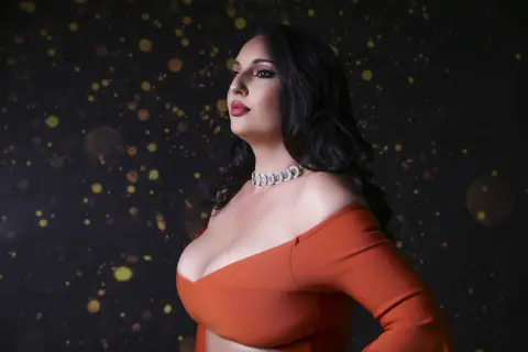 Elegant woman in an orange off-the-shoulder top and diamond necklace posing against a dark bokeh background.