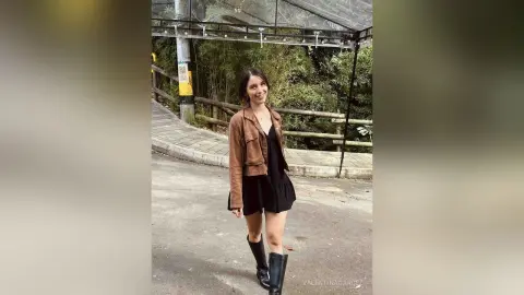 Smiling woman in a brown suede jacket and black dress walking on a paved path near lush green trees.