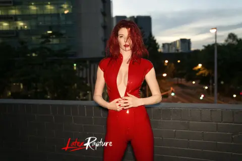 LatexRapture poses outdoors in a tight, red shiny latex bodysuit with a deep plunging neckline, showing her cleavage and red hair against a city background.