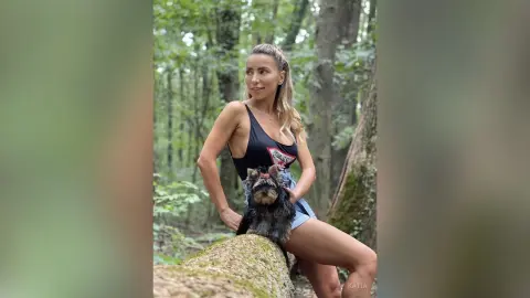 Katia poses in a black tank top and denim shorts while sitting on a log in a lush green forest with her small dog.
