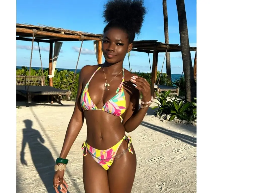 Smiling Black woman in colorful floral bikini posing on a sunny tropical beach under a wooden pergola.