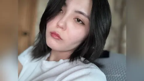 Young woman with dark hair wearing a white ribbed top in a soft, natural indoor setting.