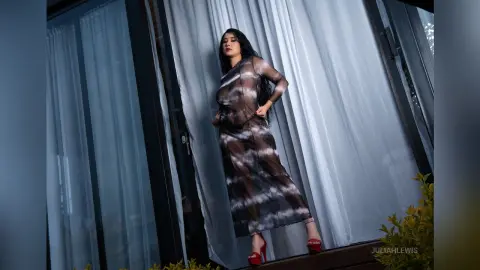 JuliahLewis stands in front of sheer white curtains wearing a long, translucent black mesh dress.