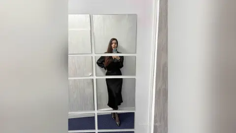 Elegant woman in a black satin dress taking a mirror selfie in a modern minimalist room.