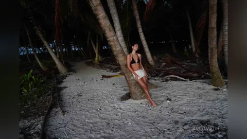 LucyMartin leans against a palm tree trunk on a sandy beach wearing a black bikini top and white bottoms.