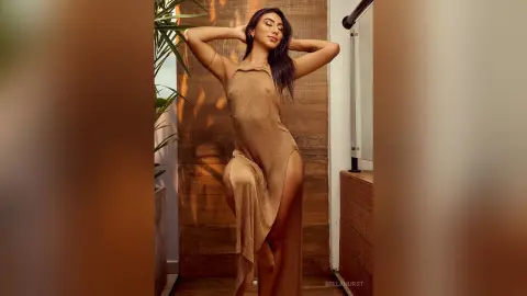Elegant woman in a shimmering gold halter dress posing in warm sunlight against a wooden wall.