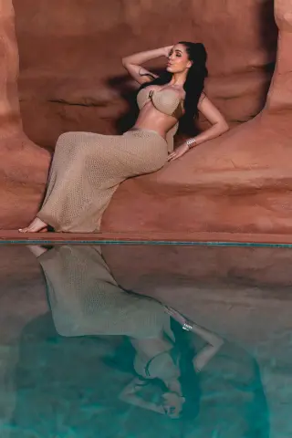 Elegant woman in a beige crochet bikini and long skirt relaxing by a desert rock pool at sunset.