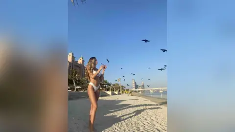 LilyReyes stands on a sandy beach in a white bikini, looking up at birds flying in a clear blue sky near a large resort building.