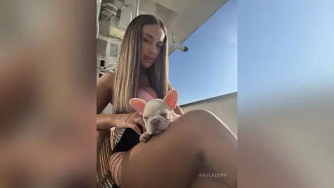 KaylaDepp poses outdoors in a pink top and black shorts, holding a small white puppy against her legs.