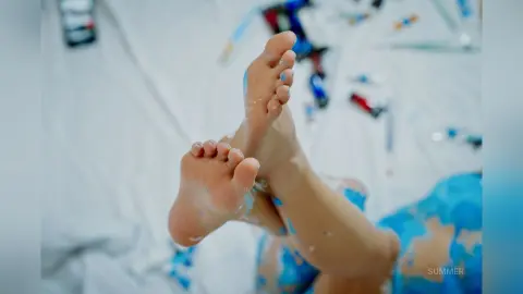Summer lifts her bare feet toward the camera, showing her toes and soles against a white bedsheet.