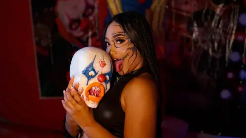 Woman in black outfit holding a creepy clown mask with spooky Halloween makeup in a dark setting.