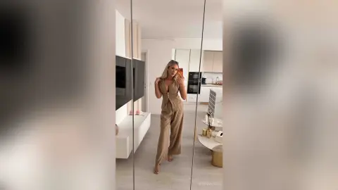Blonde woman in a stylish tan jumpsuit posing in a modern, bright minimalist living room.