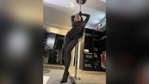 NicoleBella poses naked against a silver pole, wearing sheer black stockings and black high heels.
