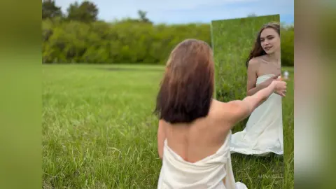 Young woman in a white strapless dress looking into a mirror in a lush green meadow during golden hour.