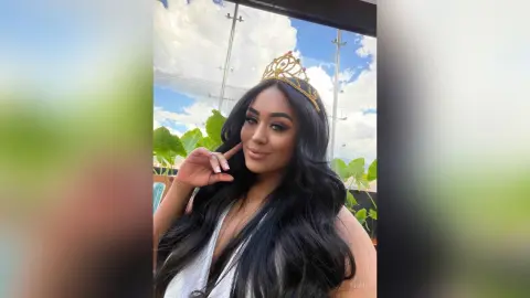 Beautiful woman with long wavy hair wearing a gold crown in a sunny outdoor garden setting.