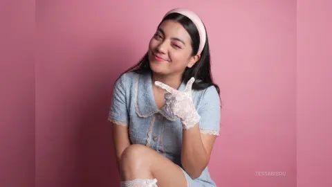 Smiling woman in a light blue vintage lace dress and white gloves posing against a pink background.