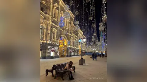 Woman sitting on a park bench at night in front of a grand, illuminated building with festive fairy lights.