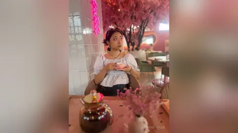 Young woman in a white off-the-shoulder top enjoying tea in a dreamy pink cafe with cherry blossom decor.