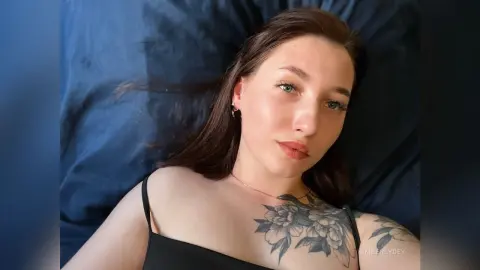 Young woman with long brown hair and floral tattoos relaxing on a blue pillow in a moody, intimate setting.