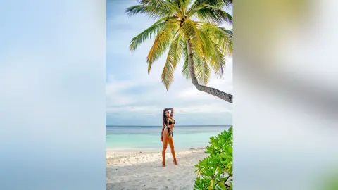 Slim woman in a black bikini standing on a sunny tropical beach under a palm tree.