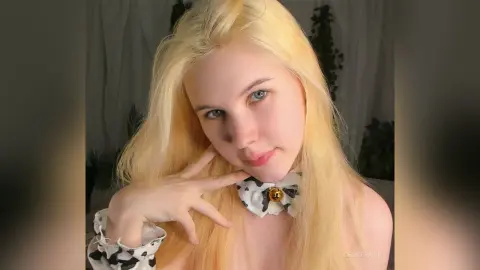 Blonde woman in a white polka dot blouse posing with a soft, dreamy expression in a bright indoor setting.