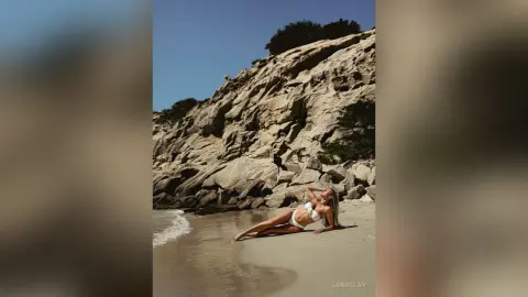 Blonde woman in a white bikini relaxing on a sunny rocky beach by the ocean waves.