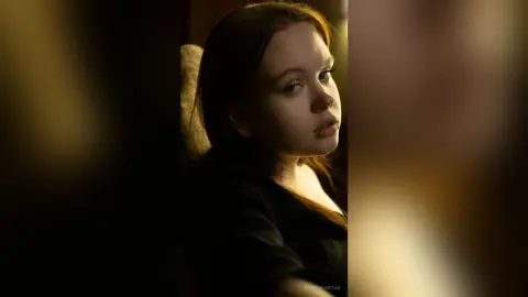 Young woman in a black top looking thoughtfully into the distance in warm, moody indoor lighting.