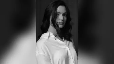 Portrait of a young woman with wavy hair wearing a white button-down shirt in a moody black and white setting.
