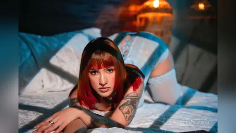 Tattooed woman with red hair in white lace lingerie lounging on a bed in moody, cinematic lighting.