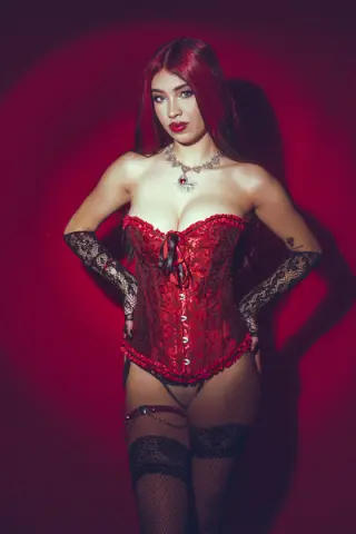 Red-haired woman in a red lace corset and black lace lingerie posing against a moody red background.