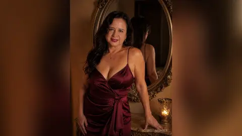 IsabellaBennet poses in a deep red satin dress with thin straps, standing next to an oval mirror and a lit candle.